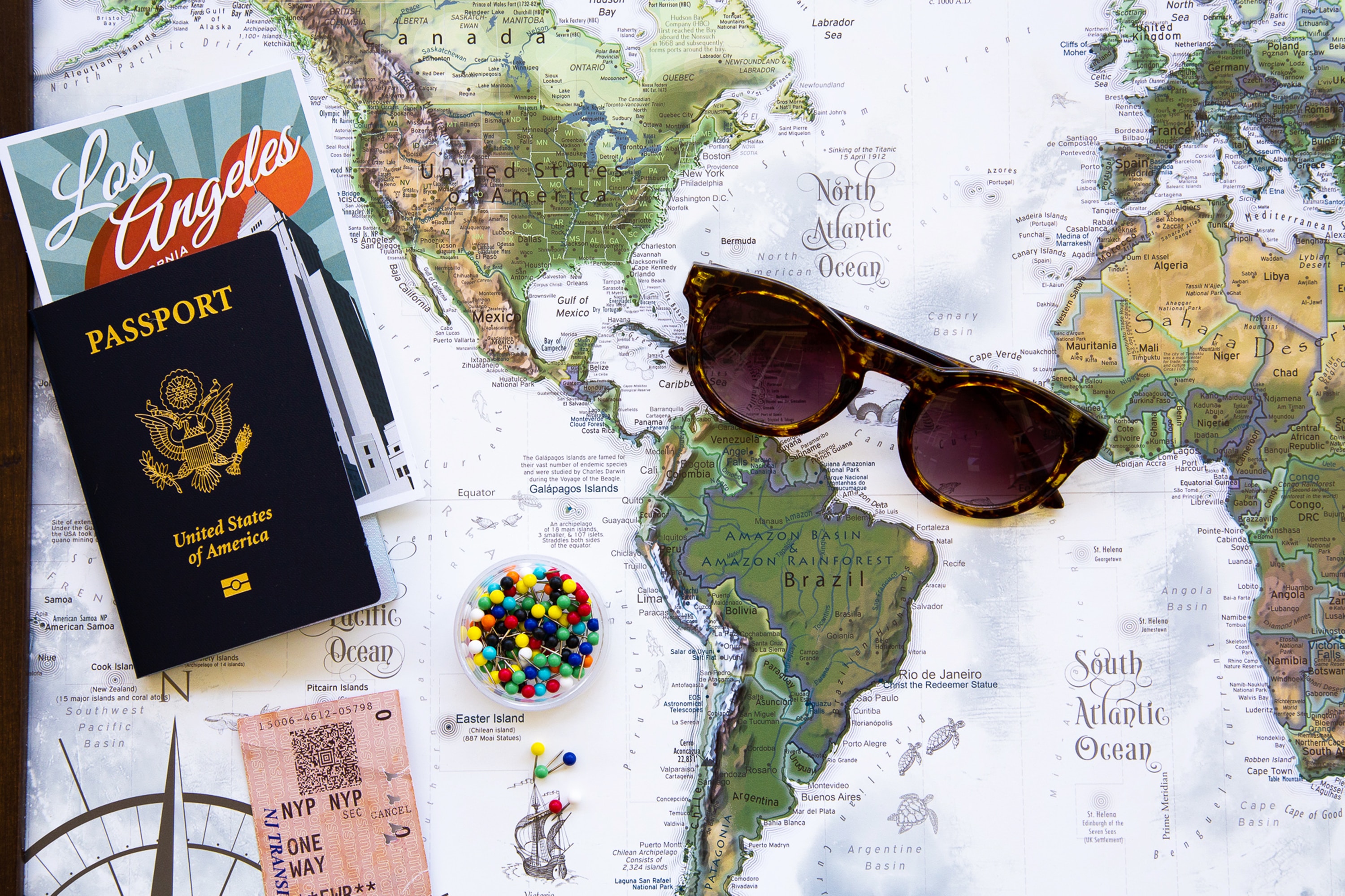 A picture of a passport with a map behind it.  Also in the picture are a part of sunglasses and a cup of pins as though a traveler is using the passport to visit places marked by pins.